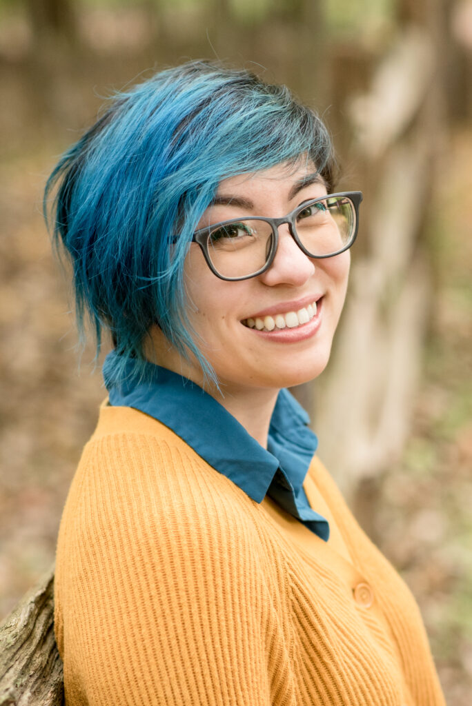 Author photo of Gale Galligan, an Asian-American cartoonist with short blue hair. They are wearing glasses, a blue collared shirt, and a yellow sweater.
