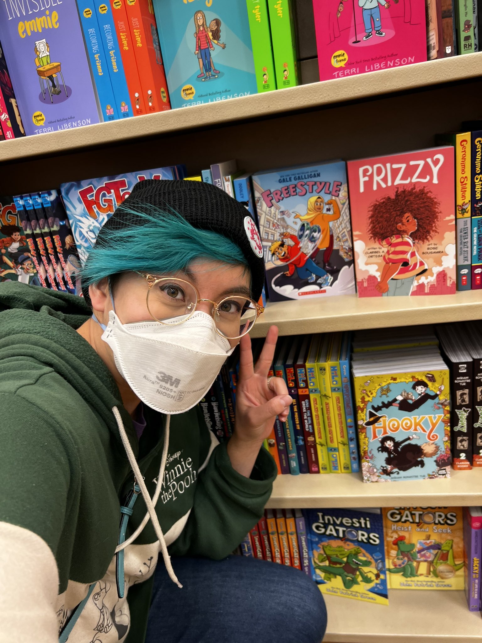 Selfie of graphic novelist Gale Galligan in front of a bookshelf featuring graphic novels FREESTYLE and FRIZZY.
